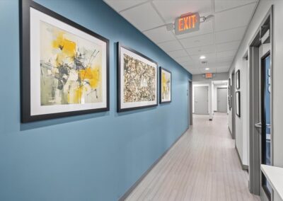 Clinical corridor at Troy Oral Surgery in Troy, Michigan, with three framed abstract prints on a slate blue wall and operatory doors down the right