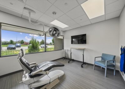 Exam room at Troy Oral Surgery in Troy, Michigan, with a black patient chair, ceiling-mounted surgical light, wall screen and a wide window onto Big Beaver Road