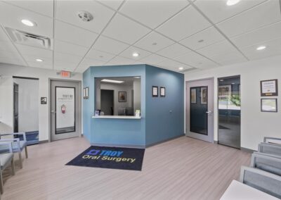 Newly constructed lobby at Troy Oral Surgery in Troy, Michigan, with a slate blue check-in window, a logo floor mat, glass doors to the waiting rooms and framed credentials on the wall