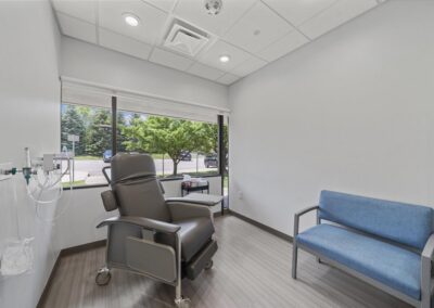 Brand new, clean recovery room at Troy Oral Surgery in Troy, Michigan, with a grey reclining recovery chair, wall-mounted oxygen and suction, a blue bench and a window onto trees