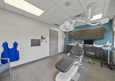 Modernly designed oral surgery suite at Troy Oral Surgery in Troy, Michigan, with a grey surgical chair, ceiling-mounted surgical light, dark wood cabinets on a slate blue wall and a lead apron on the wall