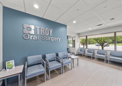 Brand new, modern waiting room at Troy Oral Surgery in Troy, Michigan, with the brushed metal practice logo on a slate blue wall, light blue chairs and windows onto the parking lot